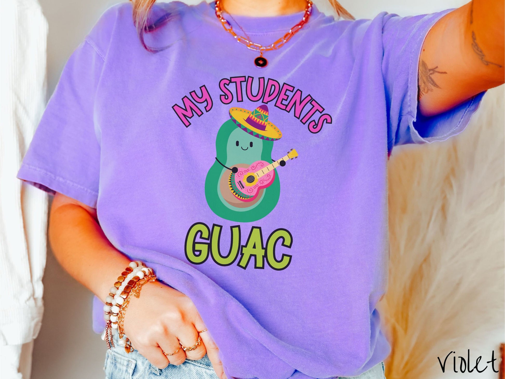 A woman wearing a cute, vintage violet colored Comfort Colors t-shirt with the text My Students Guac in pink and green font. In between the text is a smiling avocado wearing a colorful sombrero and playing a colorful mariachi guitar.