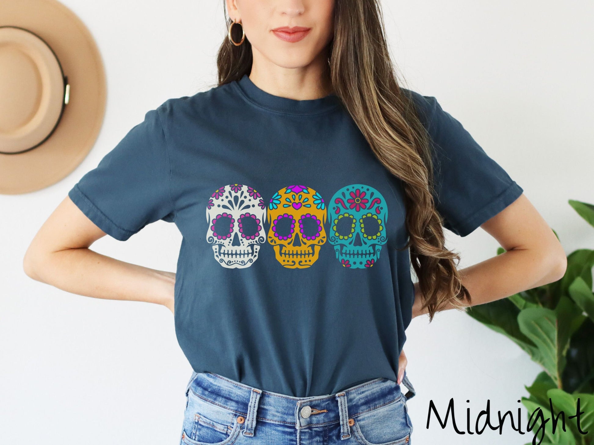 A woman wearing a cute, vintage midnight colored Comfort Colors t-shirt with three sugar skulls across the front. One is white with pink flowers, the next is orange with purple and teal flowers, and the third is teal with red flowers.