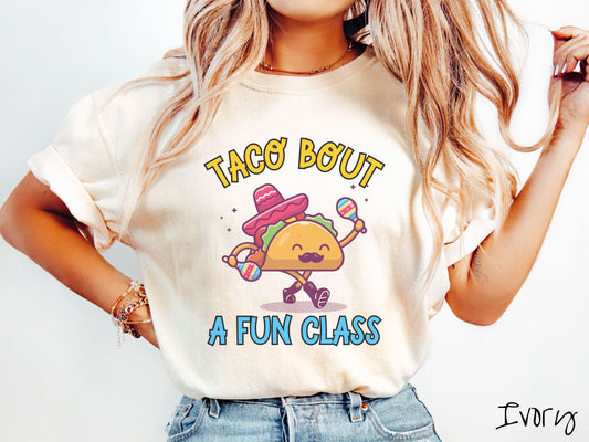 A woman wearing a cute, vintage ivory colored Comfort Colors t-shirt with text Taco Bout a Fun Class in yellow and blue font. In between the text is a smiling taco with a black mustache shaking colorful tambourines and wearing a red sombrero.