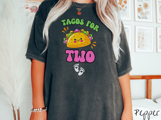 A woman wearing a cute, vintage pepper colored Comfort Colors t-shirt with text Tacos for Two in green and pink font. In between the font is a smiling taco with colorful fillings and below are two baby footprints with hearts inside.