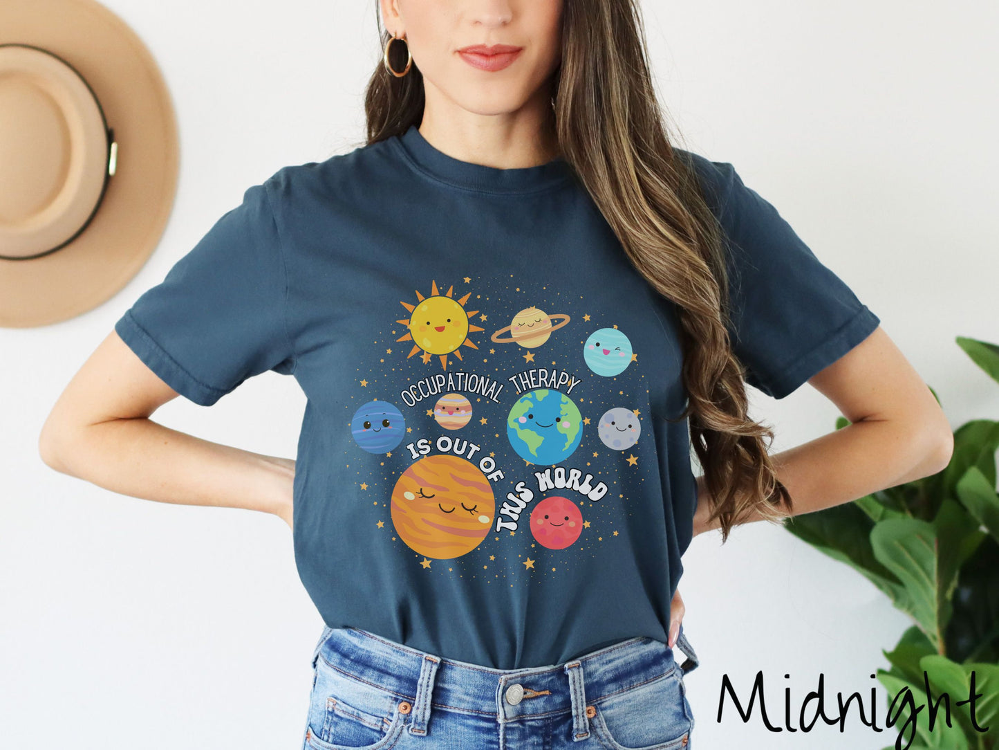 A woman wearing a midnight colored shirt showing the solar system and all the planets have smiling, happy faces. There is text saying Occupational Therapy Is Out of This World among the planets, and there are many stars twinkling in the background.