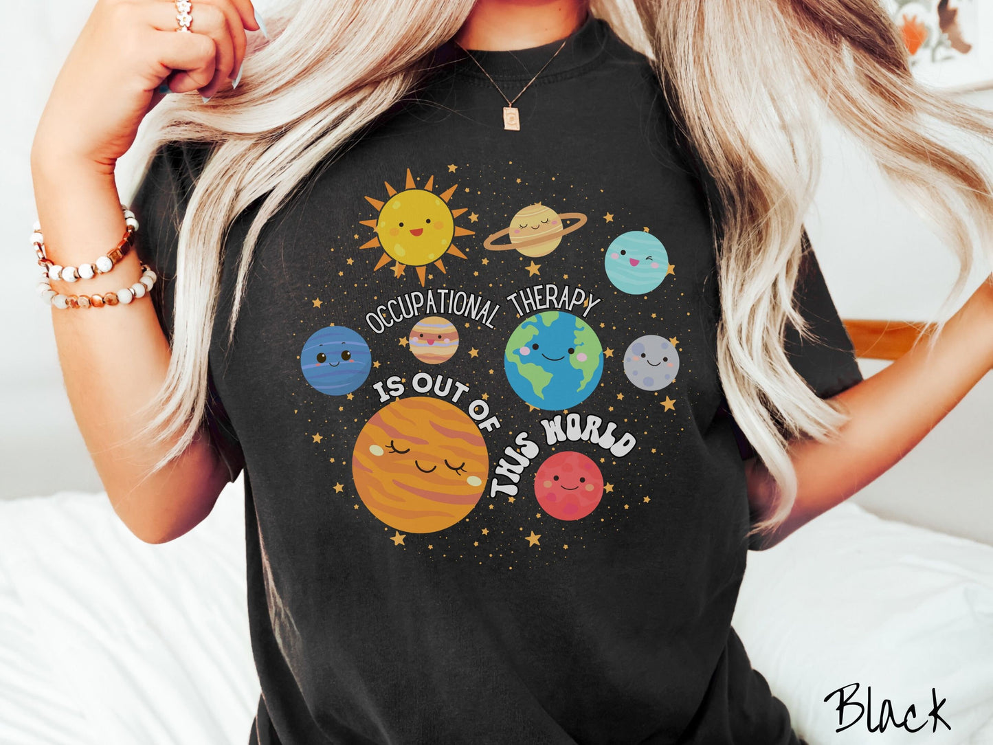 A woman wearing a black colored shirt showing the solar system and all the planets have smiling, happy faces. There is text saying Occupational Therapy Is Out of This World among the planets, and there are many stars twinkling in the background.