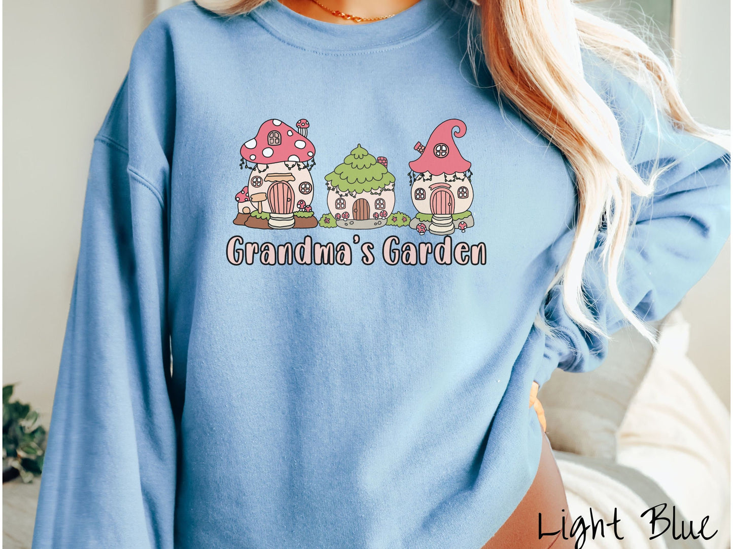 A woman wearing a cute, vintage light blue colored comfy sweatshirt with the text Grandmas Garden in light pink font. Above the text are cute little cottages for gnomes made from light and dark pink mushrooms.