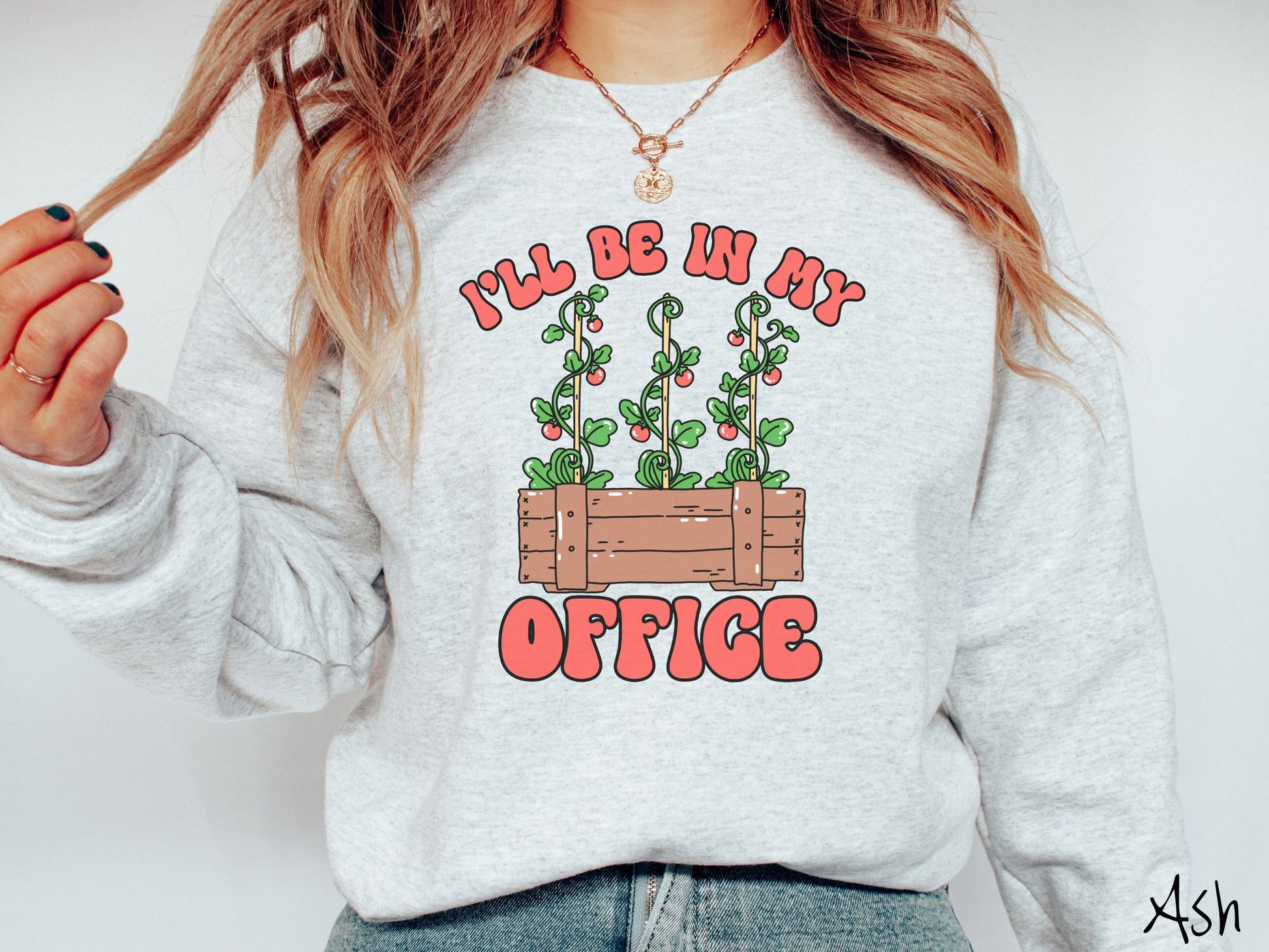 A woman wearing a cute, vintage ash colored comfy sweatshirt with the text I’ll Be in My Office in red font across the front. In between the text is a wooden plant holder with three tomato vines sprouting tomatoes climbing upwards.
