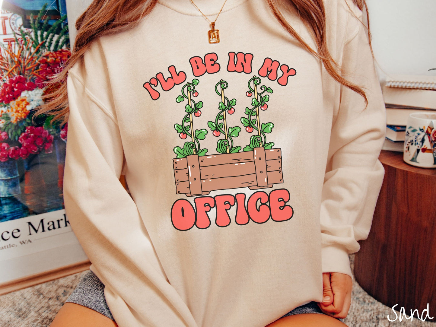 A woman wearing a cute, vintage sand colored comfy sweatshirt with the text I’ll Be in My Office in red font across the front. In between the text is a wooden plant holder with three tomato vines sprouting tomatoes climbing upwards.