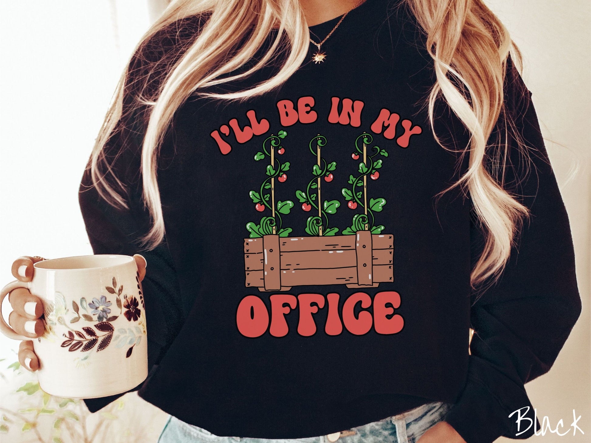 A woman wearing a cute, vintage black colored comfy sweatshirt with the text I’ll Be in My Office in red font across the front. In between the text is a wooden plant holder with three tomato vines sprouting tomatoes climbing upwards.