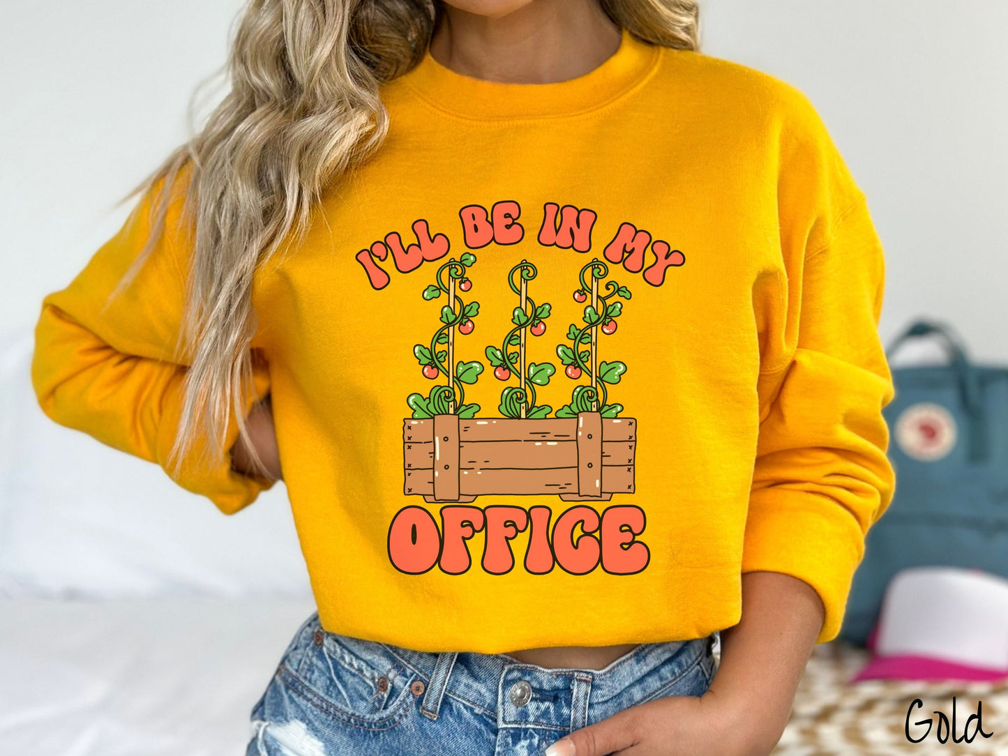 A woman wearing a cute, vintage gold colored comfy sweatshirt with the text I’ll Be in My Office in red font across the front. In between the text is a wooden plant holder with three tomato vines sprouting tomatoes climbing upwards.