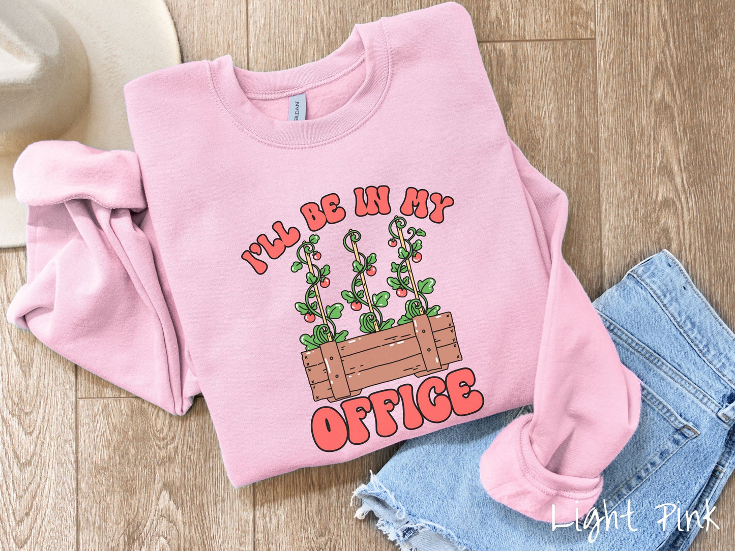 A cute, vintage light pink colored comfy sweatshirt with the text I’ll Be in My Office in red font across the front. In between the text is a wooden plant holder with three tomato vines sprouting tomatoes climbing upwards.