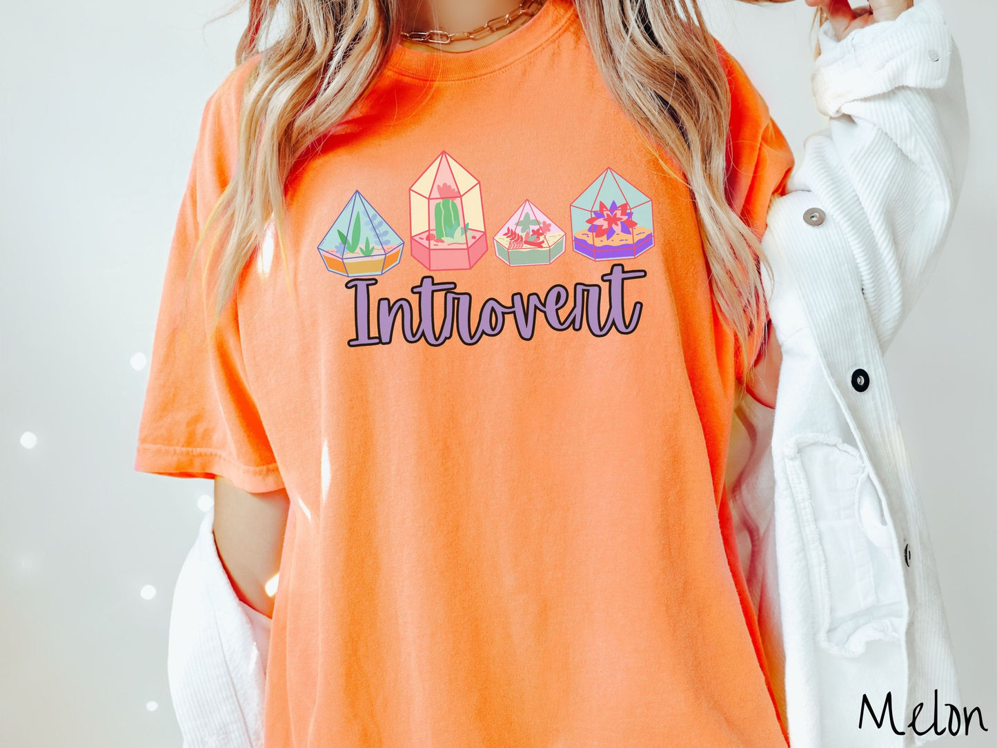 A woman wearing a cute, vintage melon colored Comfort Colors t-shirt with the text Introvert in purple font across the front. Above the text are four colorful plant terrariums with colorful flowers and cacti inside.