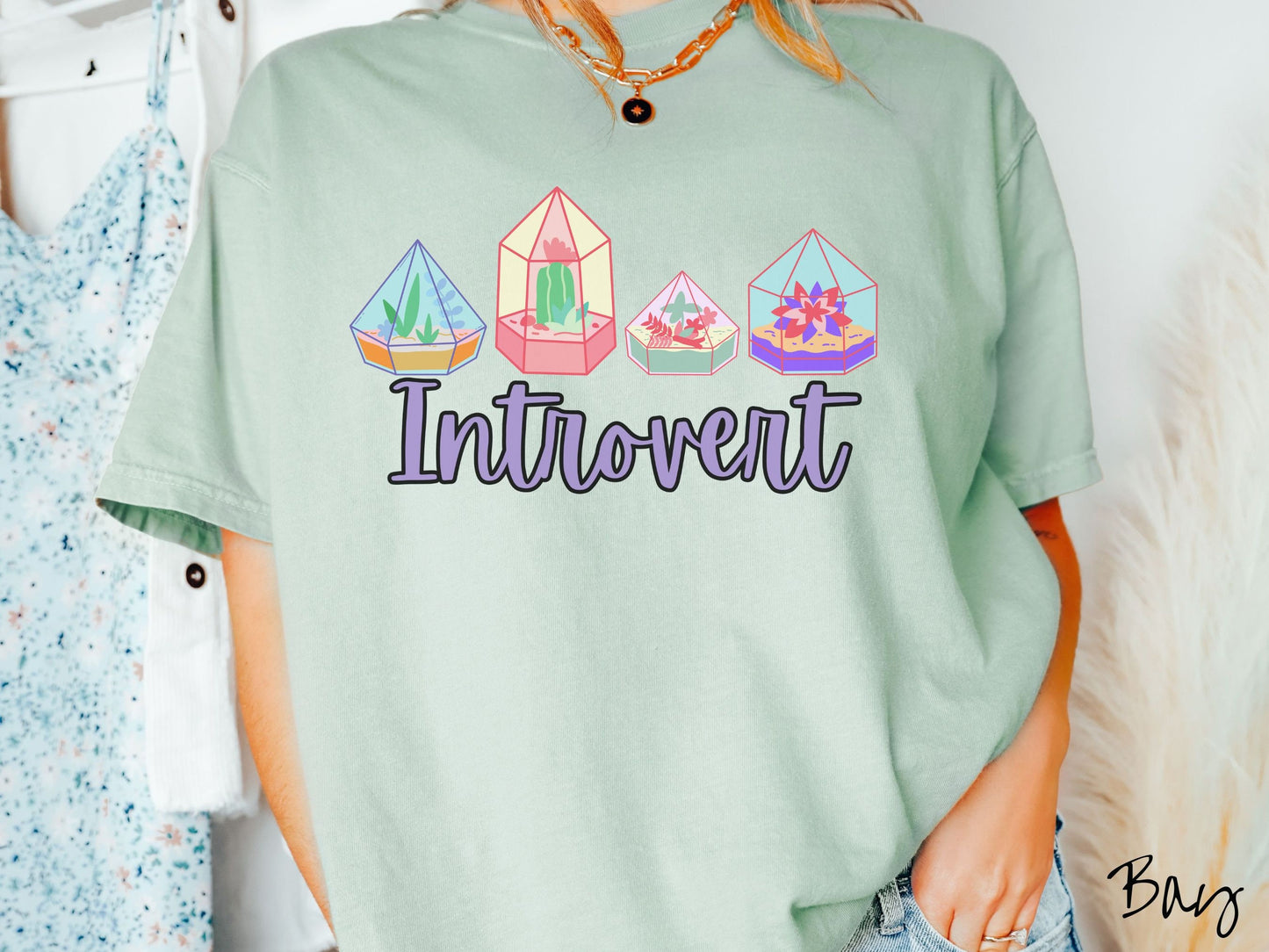 A woman wearing a cute, vintage bay colored Comfort Colors t-shirt with the text Introvert in purple font across the front. Above the text are four colorful plant terrariums with colorful flowers and cacti inside.