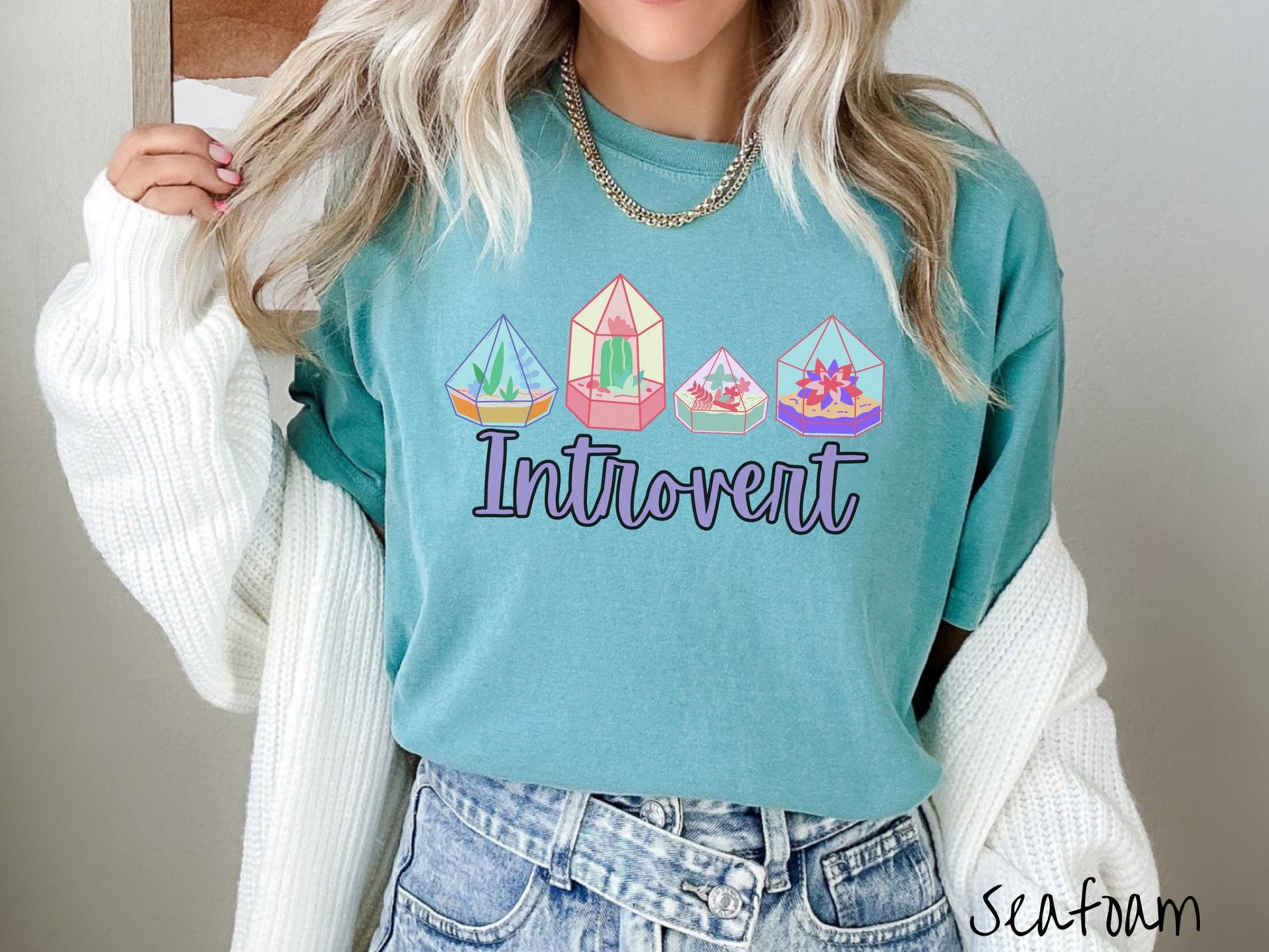 A woman wearing a cute, vintage seafoam colored Comfort Colors t-shirt with the text Introvert in purple font across the front. Above the text are four colorful plant terrariums with colorful flowers and cacti inside.