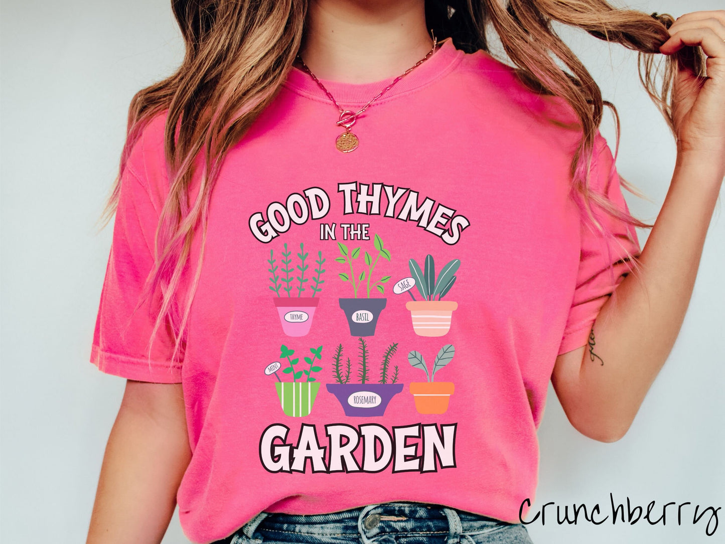 A woman wearing a cute, vintage crunchberry colored Comfort Colors t-shirt with the text Good Thymes in the Garden in white font. In between the text are different potted vegetables in colorful pots like thyme, basil, sage, and mint.