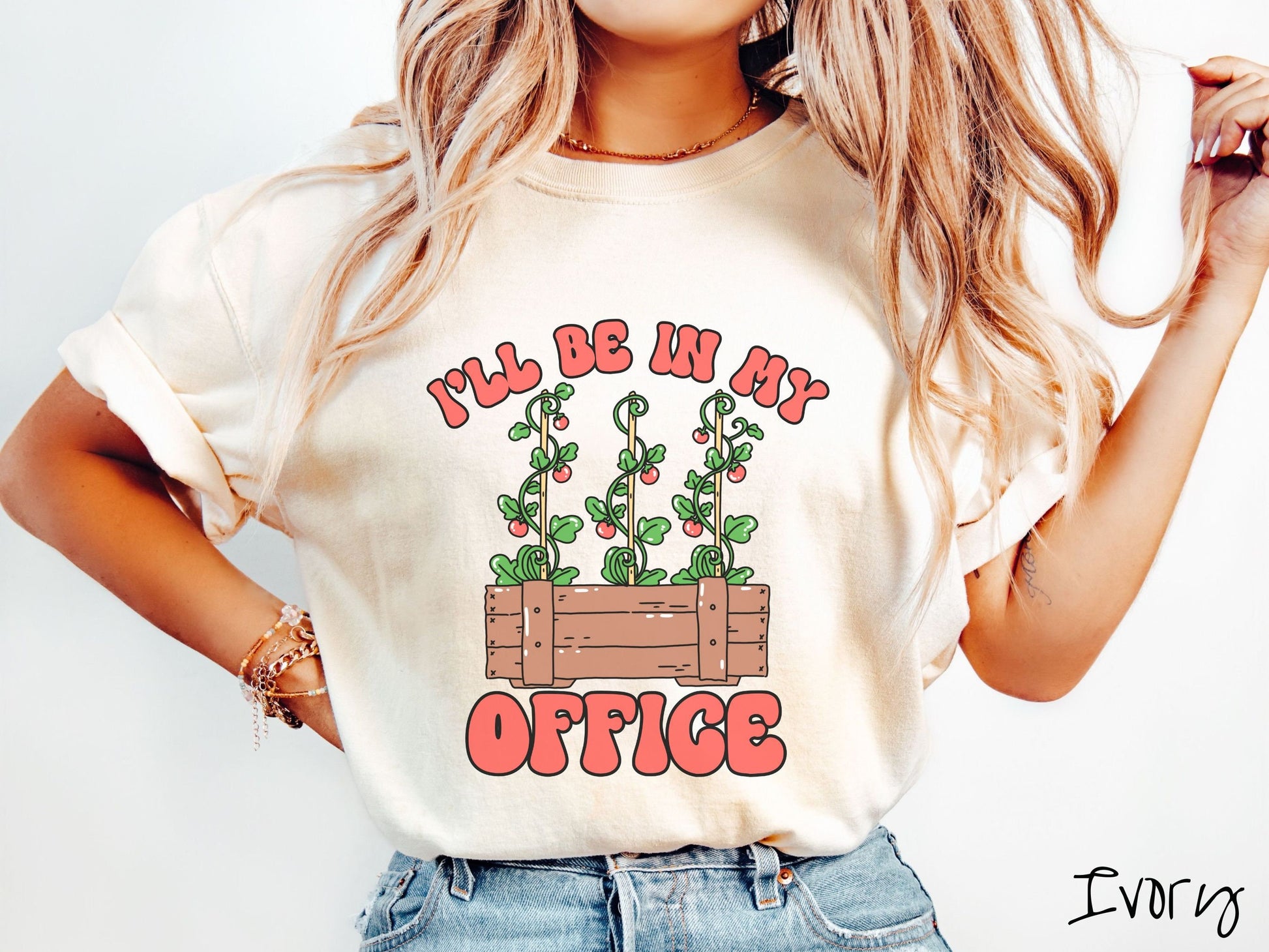 A woman wearing a cute, vintage ivory colored Comfort Colors t-shirt with the text I’ll Be in My Office in red font across the front. In between the text is a wooden plant holder with three tomato vines sprouting tomatoes climbing upwards.