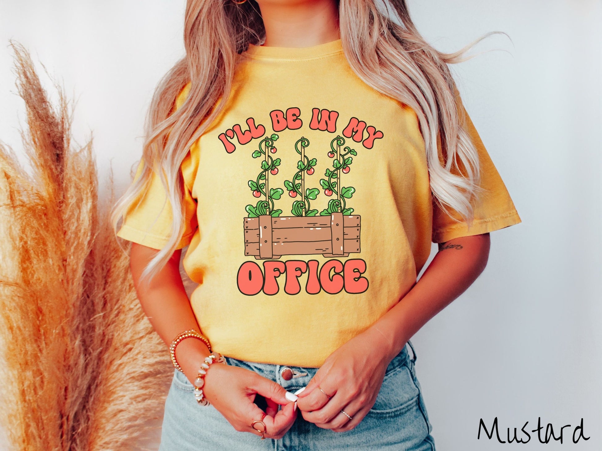 A woman wearing a cute, vintage mustard colored Comfort Colors t-shirt with the text I’ll Be in My Office in red font across the front. In between the text is a wooden plant holder with three tomato vines sprouting tomatoes climbing upwards.