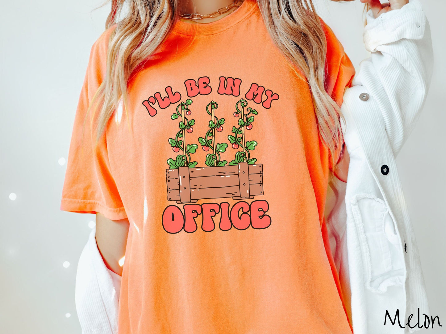 A woman wearing a cute, vintage melon colored Comfort Colors t-shirt with the text I’ll Be in My Office in red font across the front. In between the text is a wooden plant holder with three tomato vines sprouting tomatoes climbing upwards.