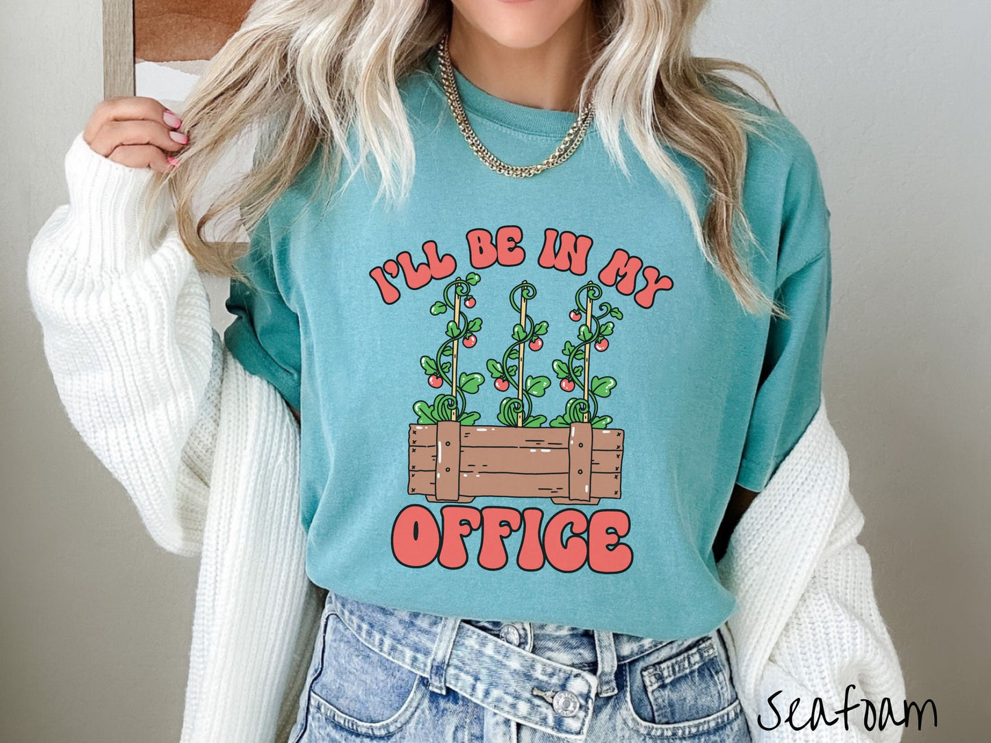 A woman wearing a cute, vintage seafoam colored Comfort Colors t-shirt with the text I’ll Be in My Office in red font across the front. In between the text is a wooden plant holder with three tomato vines sprouting tomatoes climbing upwards.