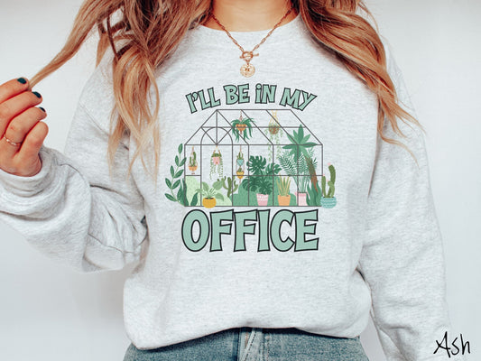 A woman wearing a cute, vintage ash colored comfy sweatshirt with the text Ill Be in My Office in green font across the front. In between the text is a glass greenhouse with all kinds of plants and flowers inside and hanging potted plants.