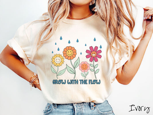 A woman wearing a cute, vintage ivory colored Comfort Colors t-shirt with the text Grow with the Flow in blue font across the bottom. Above the text are smiling yellow, orange, pink, and red flowers with raindrops falling in the background.