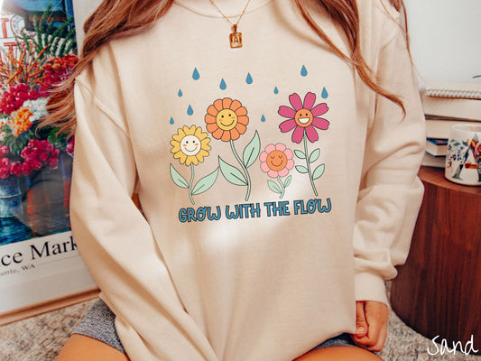 A woman wearing a cute, vintage sand colored comfy sweatshirt with the text Grow with the Flow in blue font across the bottom. Above the text are smiling yellow, orange, pink, and red flowers with raindrops falling in the background.