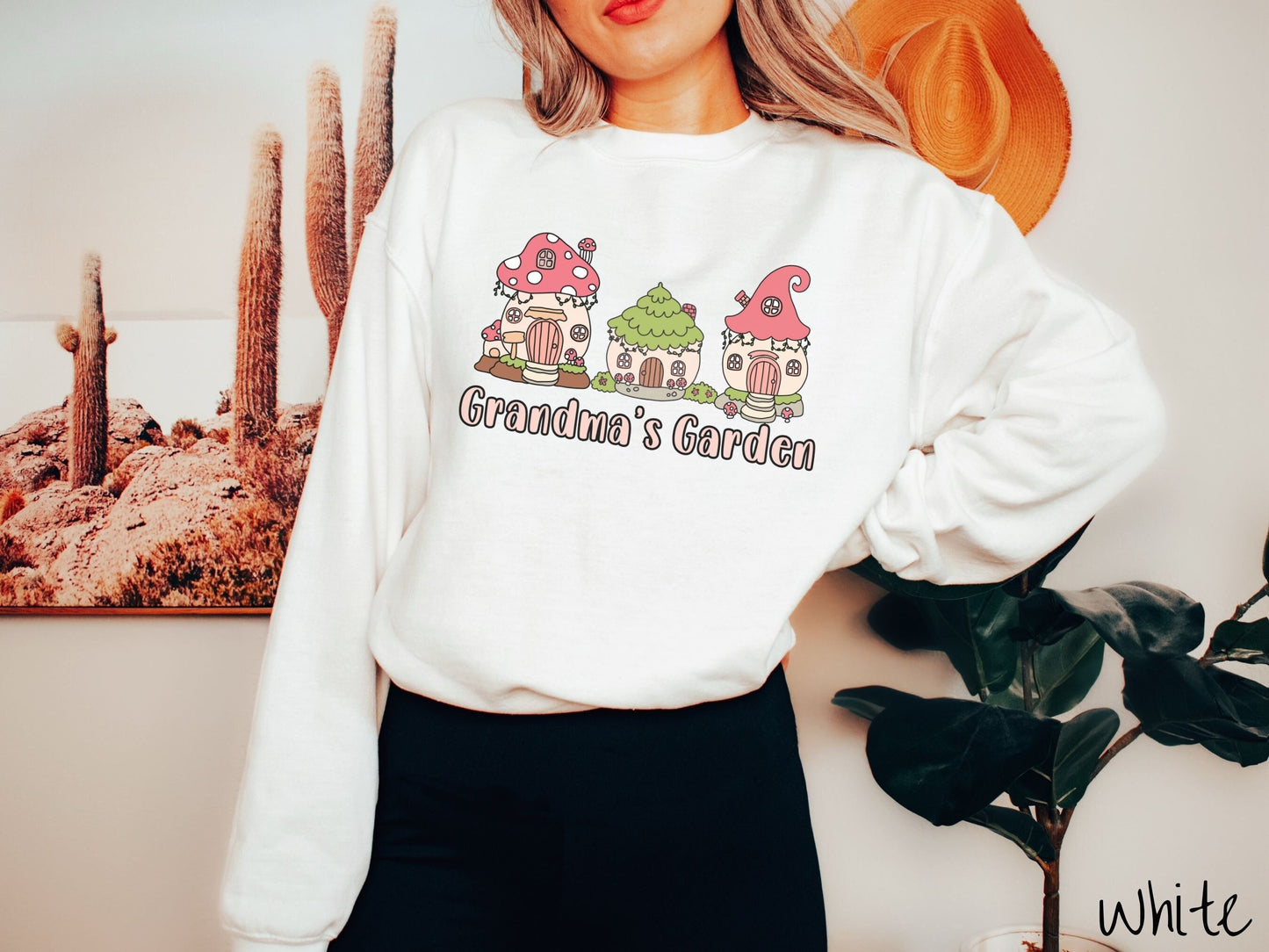 A woman wearing a cute, vintage white colored comfy sweatshirt with the text Grandmas Garden in light pink font. Above the text are cute little cottages for gnomes made from light and dark pink mushrooms.