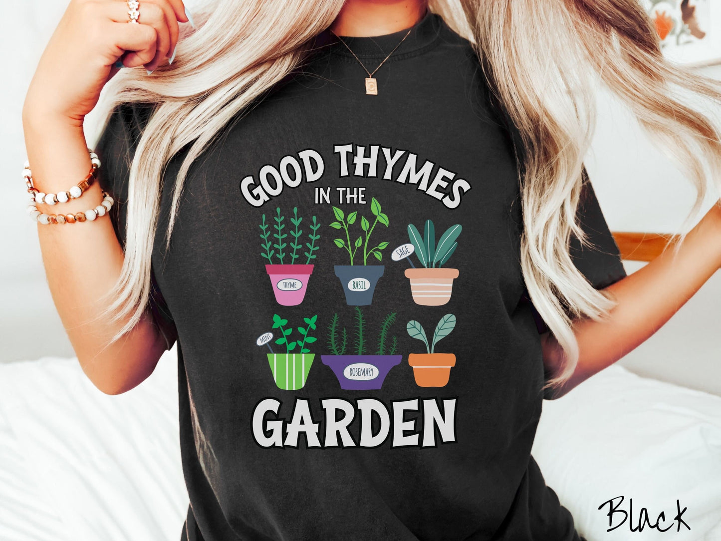 A woman wearing a cute, vintage black colored Comfort Colors t-shirt with the text Good Thymes in the Garden in white font. In between the text are different potted vegetables in colorful pots like thyme, basil, sage, and mint.