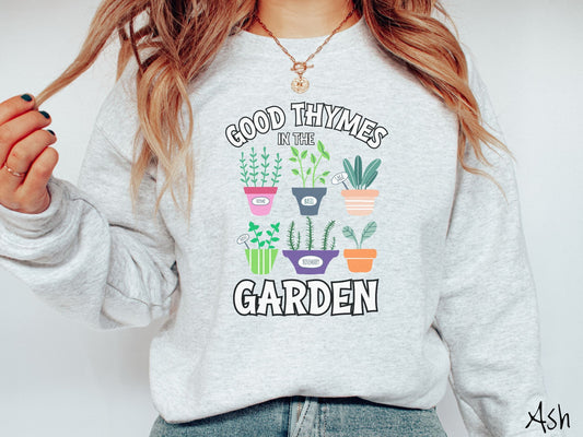 A woman wearing a cute, vintage ash colored comfy sweatshirt with the text Good Thymes in the Garden in white font. In between the text are different potted vegetables in colorful pots like thyme, basil, sage, and mint.