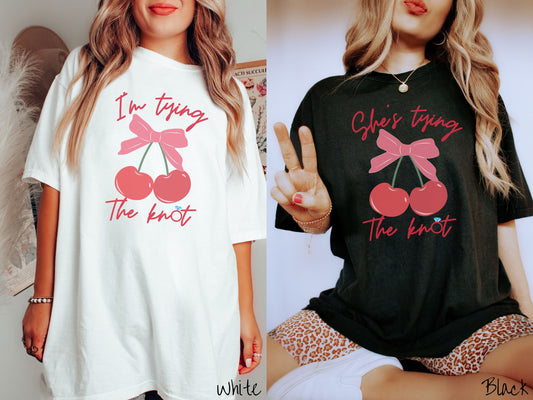 Two women wearing cute, vintage white and black colored Comfort Colors t-shirts with text Im tying the knot in red, cursive font. The o in knot is a diamond wedding ring. Between the text are two cherries tied together with a red bow.