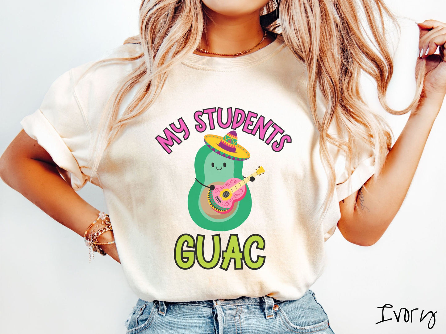 A woman wearing a cute, vintage ivory colored Comfort Colors t-shirt with the text My Students Guac in pink and green font. In between the text is a smiling avocado wearing a colorful sombrero and playing a colorful mariachi guitar.