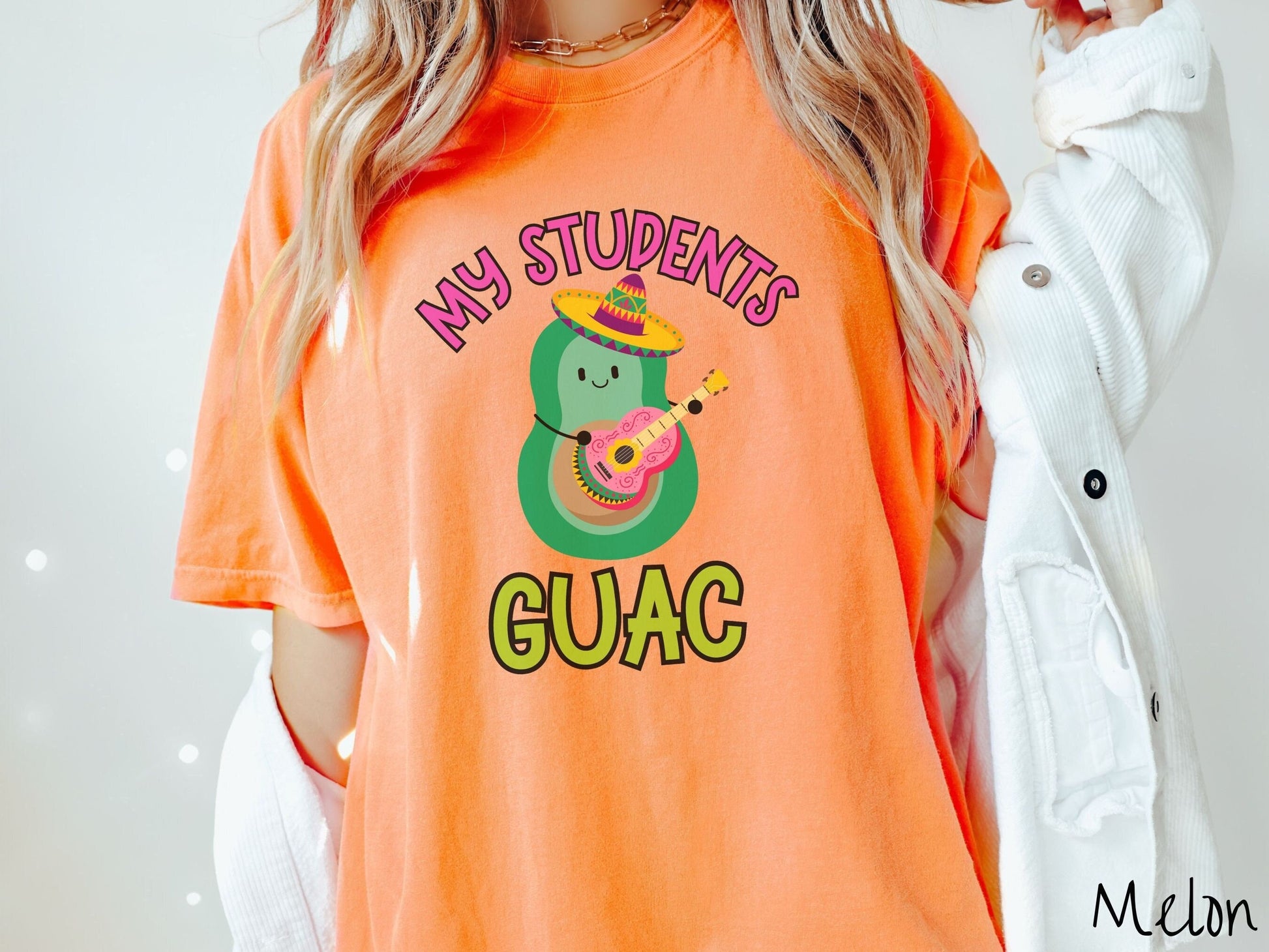 A woman wearing a cute, vintage melon colored Comfort Colors t-shirt with the text My Students Guac in pink and green font. In between the text is a smiling avocado wearing a colorful sombrero and playing a colorful mariachi guitar.