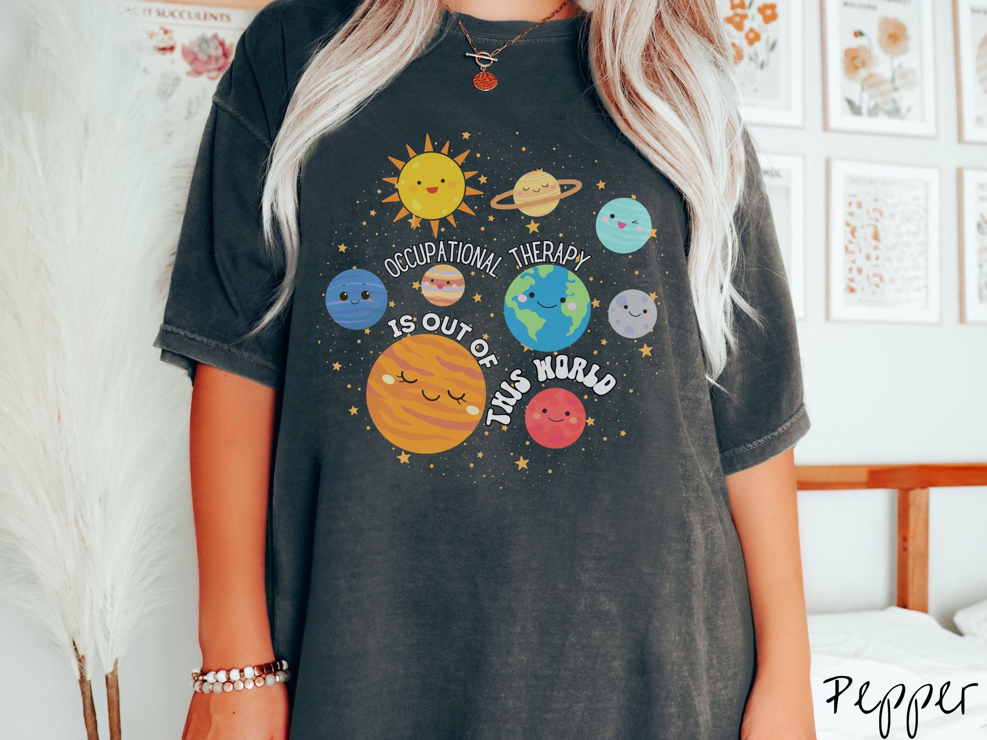 A woman wearing a pepper colored shirt showing the solar system and all the planets have smiling, happy faces. There is text saying Occupational Therapy Is Out of This World among the planets, and there are many stars twinkling in the background.