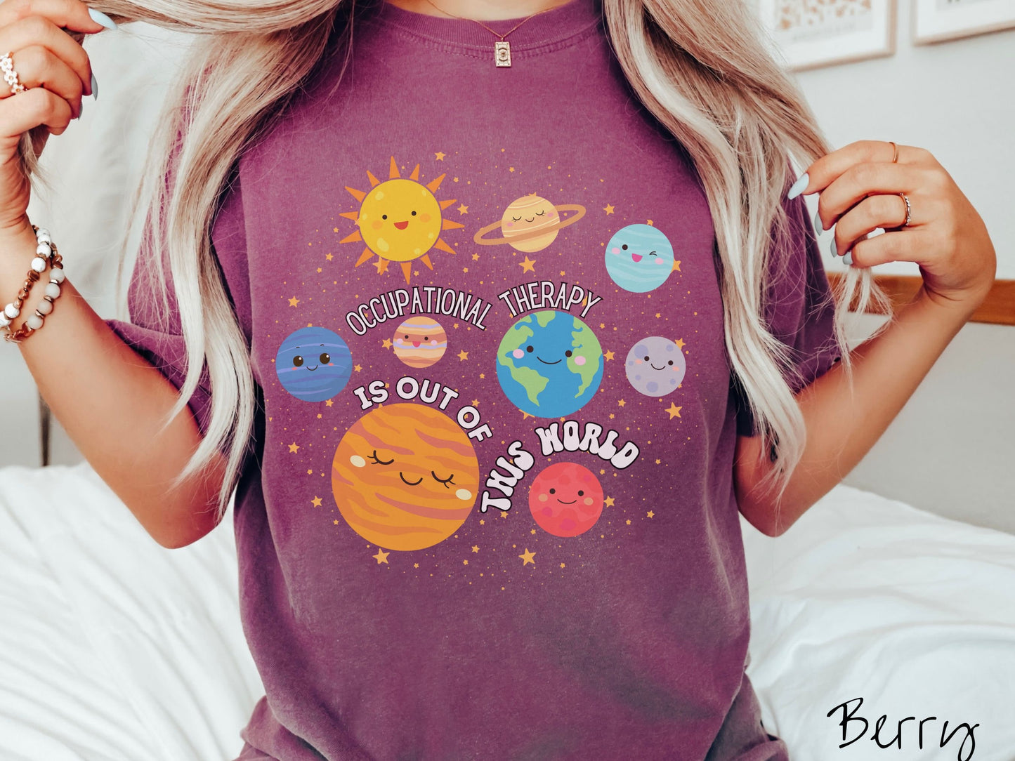 A woman wearing a berry colored shirt showing the solar system and all the planets have smiling, happy faces. There is text saying Occupational Therapy Is Out of This World among the planets, and there are many stars twinkling in the background.