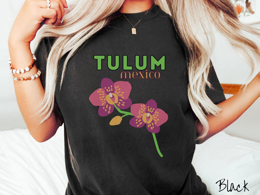 A woman wearing a vintage, black colored shirt with the text Tulum Mexico in green and yellow font, respectively, at the top and below it are two beautiful pink and purple flowers with green stems.