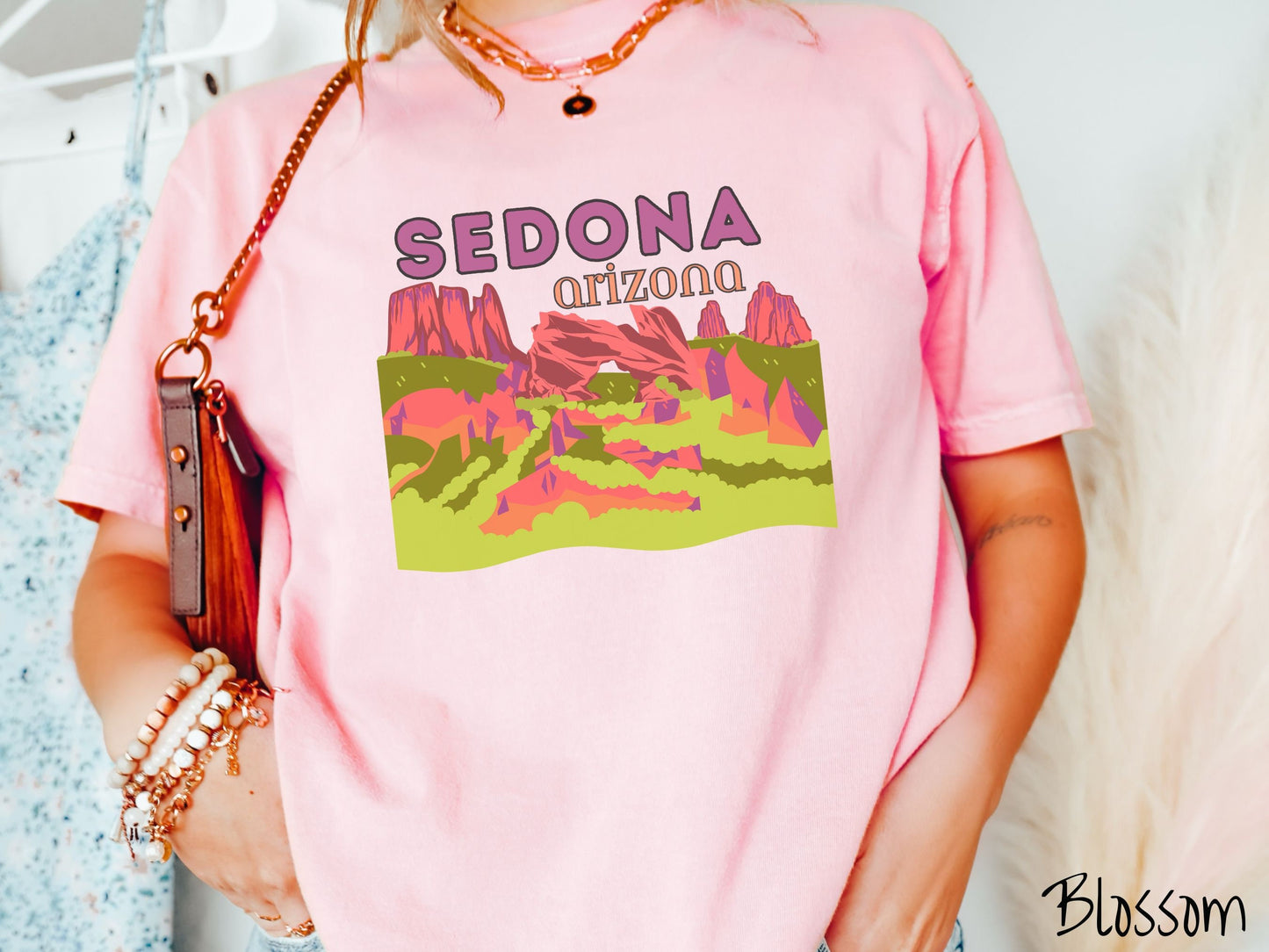 A woman wearing a vintage, blossom colored comfort colors t-shirt with the text Sedona Arizona in purple and orange font, respectively, and below that is a picture of red rock canyons and green meadows.