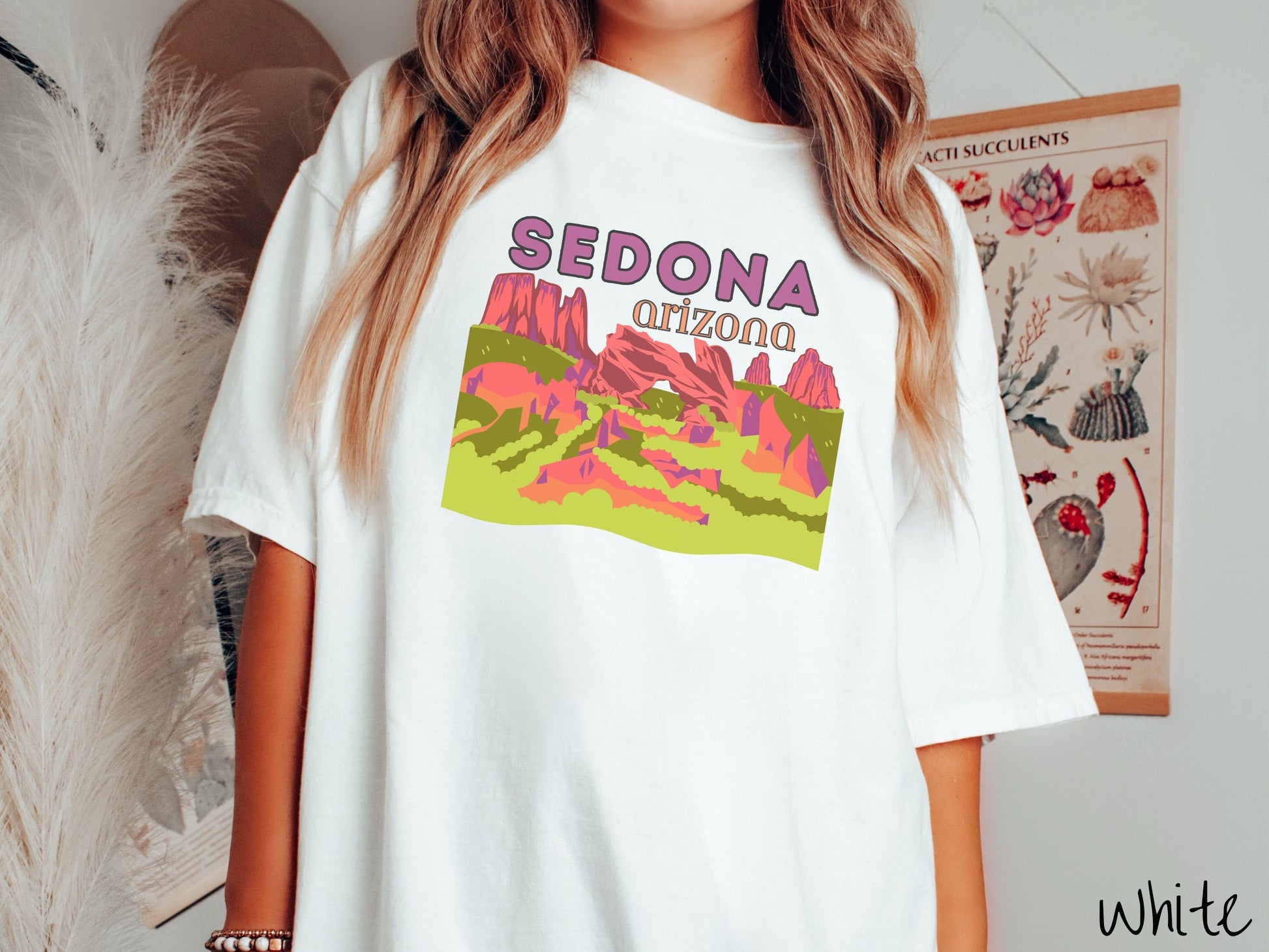 A woman wearing a vintage, white colored comfort colors t-shirt with the text Sedona Arizona in purple and orange font, respectively, and below that is a picture of red rock canyons and green meadows.