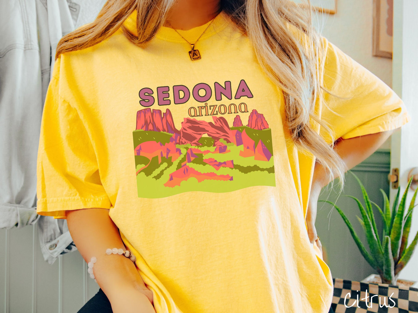 A woman wearing a vintage, citrus colored comfort colors t-shirt with the text Sedona Arizona in purple and orange font, respectively, and below that is a picture of red rock canyons and green meadows.