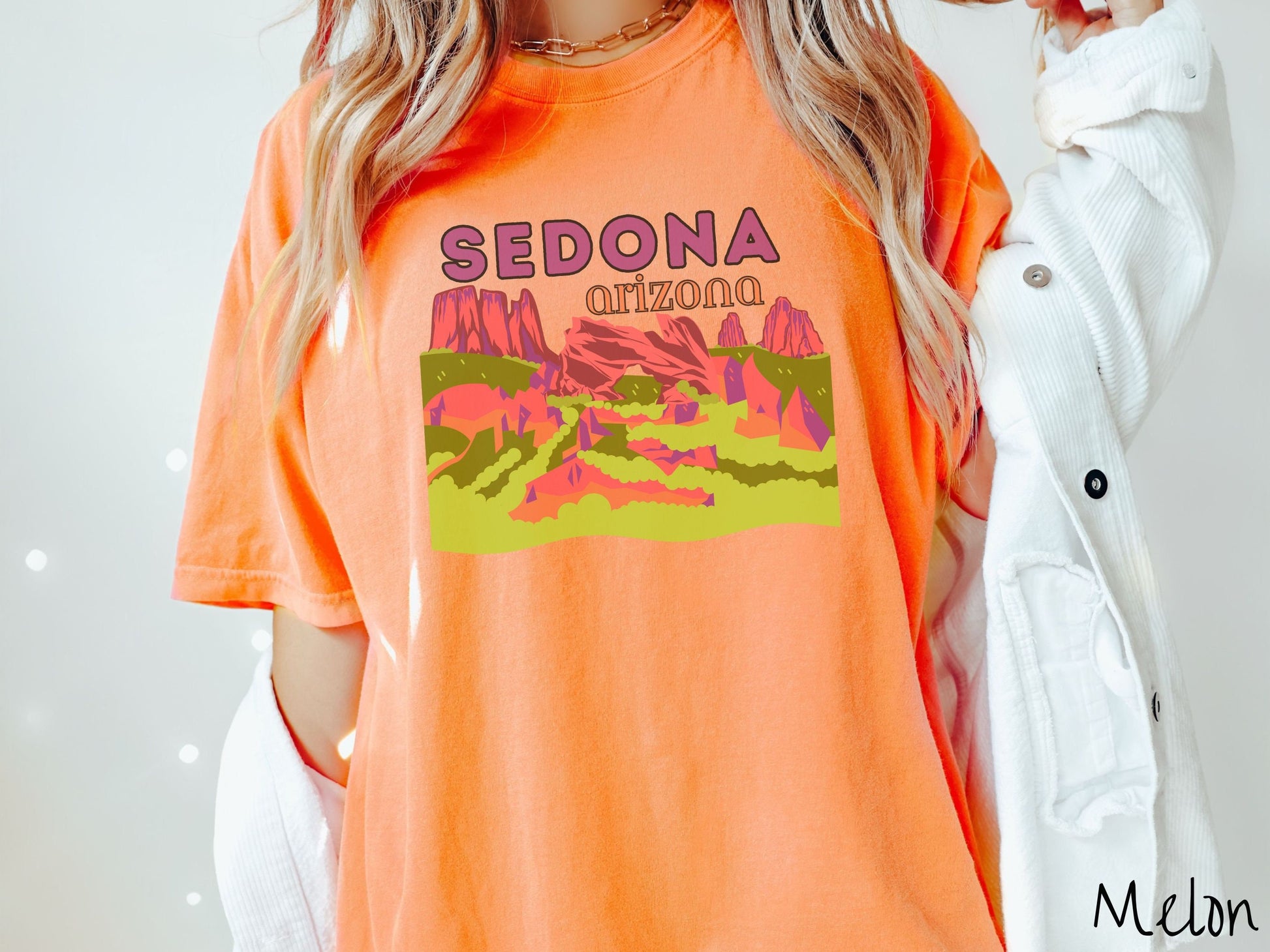 A woman wearing a vintage, melon colored comfort colors t-shirt with the text Sedona Arizona in purple and orange font, respectively, and below that is a picture of red rock canyons and green meadows.