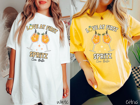 Two women wearing cute, vintage white and citrus colored Comfort Colors t-shirts with text Love at First Spritz in yellow font, the O in Love is a diamond wedding ring. Between the text are two cocktail glasses of aperol spritz toasting.