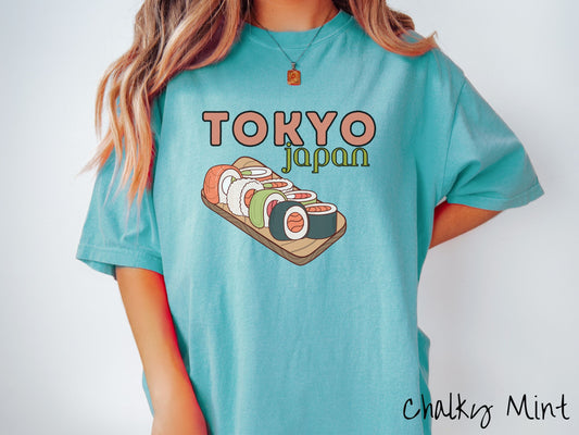 A woman wearing a vintage, chalky mint colored shirt with the text Tokyo Japan across the top in salmon and green font, respectively, and below is an assortment of colorful sushi rolls on a narrow wooden plate.