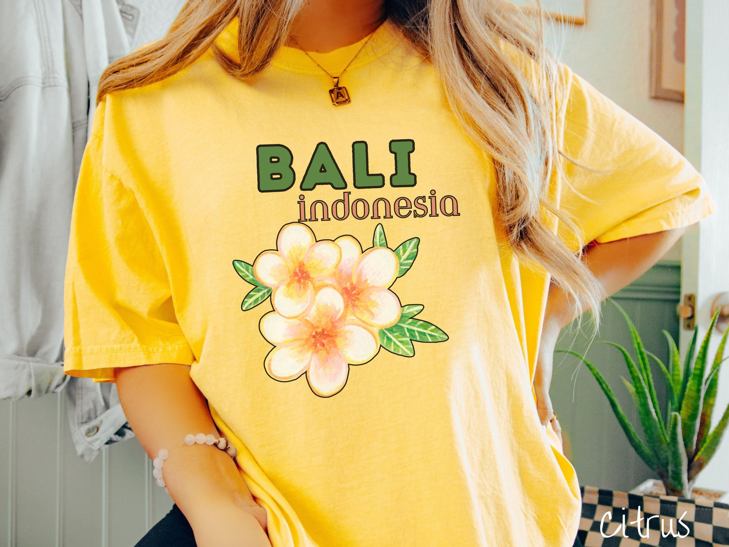 A woman wearing a vintage, citrus colored shirt with the text Bali Indonesia in green and yellow font, respectively, and a picture underneath of three beautiful yellow and orange flowers with green leaves.