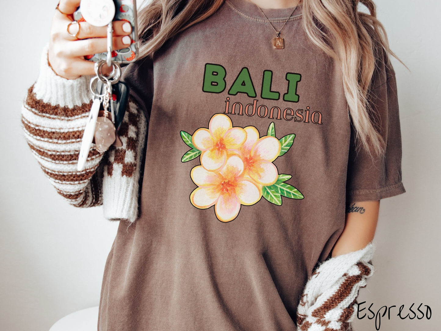A woman wearing a vintage, espresso colored shirt with the text Bali Indonesia in green and yellow font, respectively, and a picture underneath of three beautiful yellow and orange flowers with green leaves.