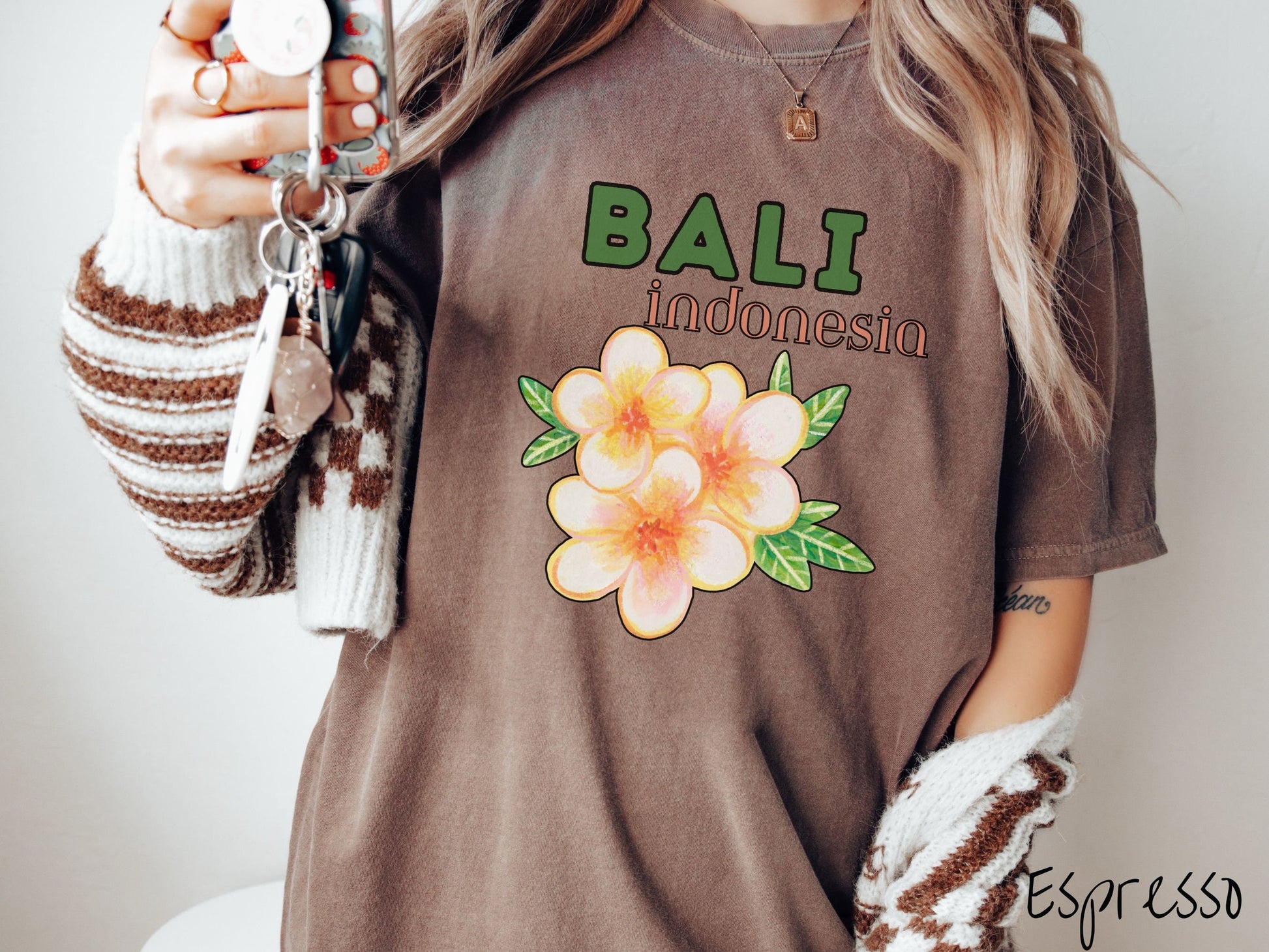 A woman wearing a vintage, espresso colored shirt with the text Bali Indonesia in green and yellow font, respectively, and a picture underneath of three beautiful yellow and orange flowers with green leaves.