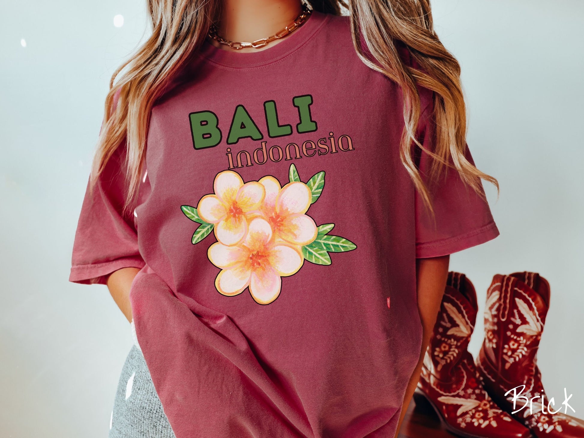 A woman wearing a vintage, brick colored shirt with the text Bali Indonesia in green and yellow font, respectively, and a picture underneath of three beautiful yellow and orange flowers with green leaves.