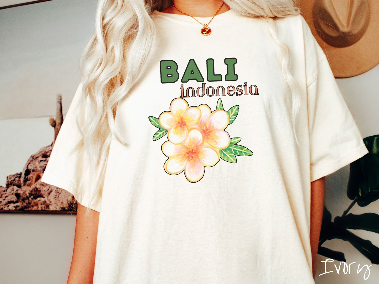 A woman wearing a vintage, ivory colored shirt with the text Bali Indonesia in green and yellow font, respectively, and a picture underneath of three beautiful yellow and orange flowers with green leaves.