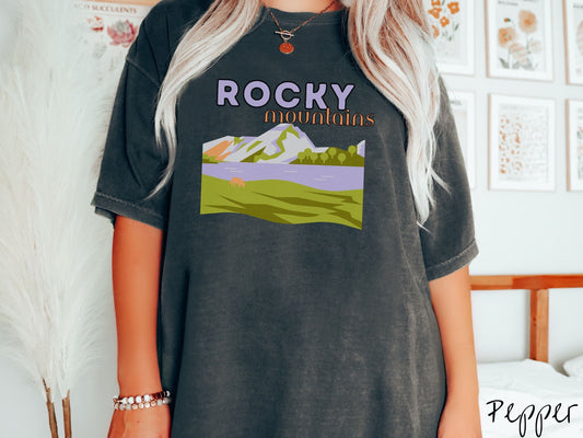 A woman wearing a vintage, pepper colored shirt with the text Rocky Mountains at the top and below is a scene of a massive river running beneath snowy mountains in the distance, a lone brown cow can be seen grazing in the grass next to the water.