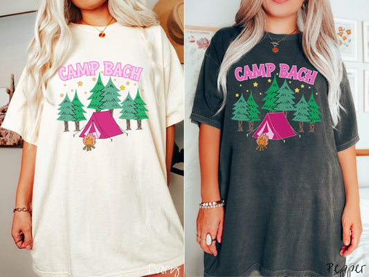 Two women wearing cute, vintage ivory and pepper colored Comfort Colors t-shirts with text Camp Bach in pink, uppercase font. Below the text is a pink tent in between green trees and in front of a campfire with stars in the sky above the trees.