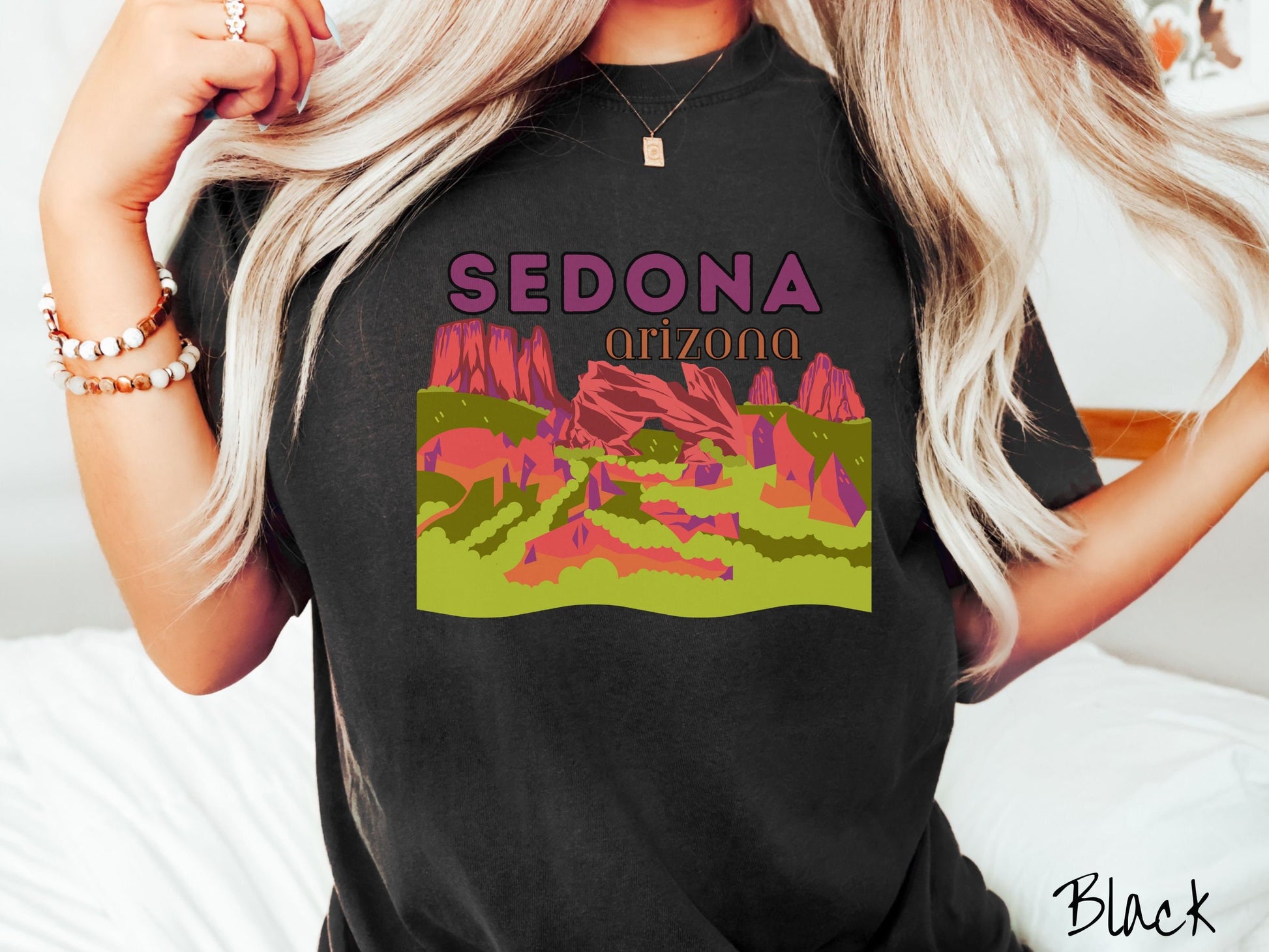 A woman wearing a vintage, black colored comfort colors t-shirt with the text Sedona Arizona in purple and orange font, respectively, and below that is a picture of red rock canyons and green meadows.