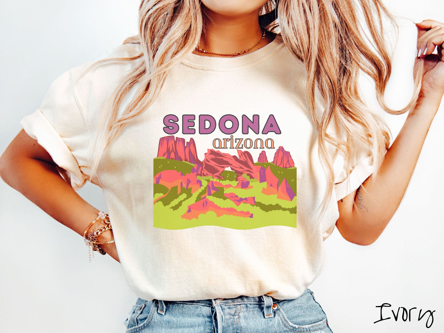 A woman wearing a vintage, ivory colored comfort colors t-shirt with the text Sedona Arizona in purple and orange font, respectively, and below that is a picture of red rock canyons and green meadows.