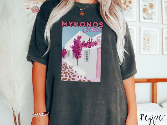 A woman wearing a vintage, pepper colored shirt with the text Mykonos Greece at the top, and beneath is a picture of Cycladic architecture, a beautiful white home with purple trees in the front yard, and a pink driveway with brown stone.