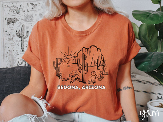 A woman wearing a cute, vintage yam colored Comfort Colors t-shirt with text Sedona, Arizona in white font. Above is a black outline of a desert landscape with the sun and mountains in the background, and cacti and rocks in the foreground.