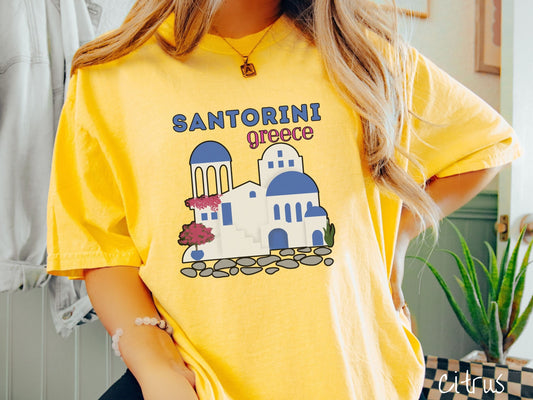 A woman wearing a cute, vintage citrus colored Comfort Colors t-shirt with text Santorini Greece in blue and pink font. Below that is a picture of the iconic cave houses with white walls and blue trim, with cobble-stone roads and red trees.